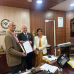 Prof. (Vd.) P.K. Prajapati and Shri Segar Sampathkumar shaking hands across a conference table, flanked by the official AIIA and GIC delegation under portraits of Indian leadership.