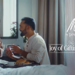 A romantic lifestyle image for Mia by Tanishq's Joy of Gifting Festival featuring a man surprising a woman with a jewelry gift box during breakfast in bed.