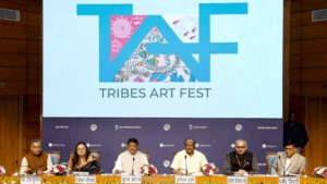 A panel of six officials seated at a press conference table under a large screen displaying the "TAF TRIBES ART FEST" logo...
