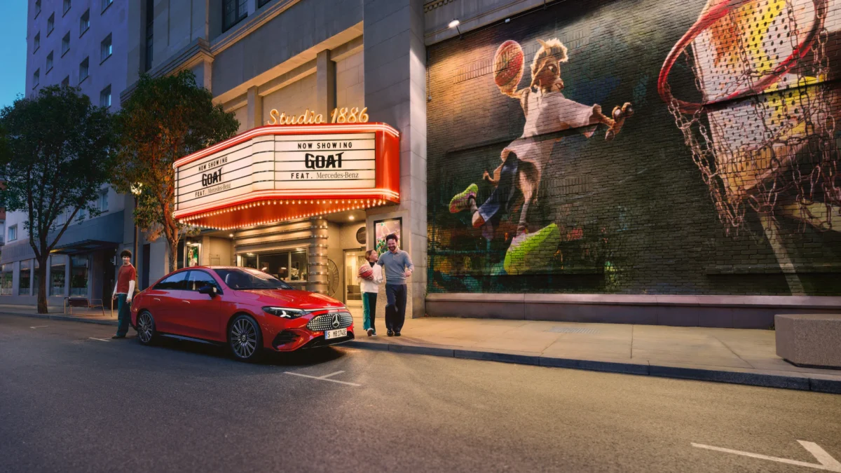 Side profile of the red Mercedes-Benz CLA EV parked in front of 'Studio 1886' cinema with 'GOAT' featured on the marquee, showcasing the entry-level luxury electric sedan's sleek aerodynamic design.
