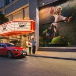 Side profile of the red Mercedes-Benz CLA EV parked in front of 'Studio 1886' cinema with 'GOAT' featured on the marquee, showcasing the entry-level luxury electric sedan's sleek aerodynamic design.