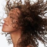 A high-resolution image of a woman with voluminous, curly brown hair blowing in the wind against a white background. This visual represents Hindustan Unilever's (HUL) 2026 strategic shift toward "Scalp-First Thinking" and microbiome-focused hair health.
