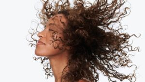 A high-resolution image of a woman with voluminous, curly brown hair blowing in the wind against a white background. This ...
