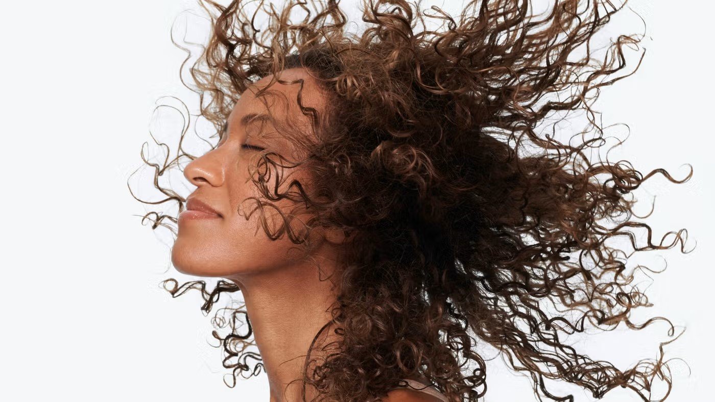 A high-resolution image of a woman with voluminous, curly brown hair blowing in the wind against a white background. This visual represents Hindustan Unilever's (HUL) 2026 strategic shift toward "Scalp-First Thinking" and microbiome-focused hair health.