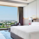 A bright, modern luxury bedroom at the Indore Marriott Hotel in India featuring floor-to-ceiling windows with a wide city view, a plush king-sized bed, and elegant minimalist decor.