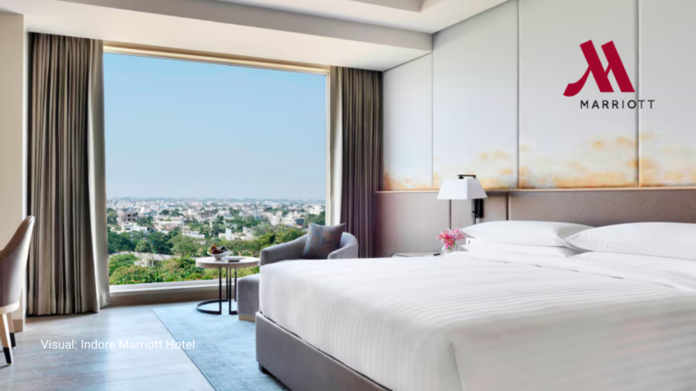A bright, modern luxury bedroom at the Indore Marriott Hotel in India featuring floor-to-ceiling windows with a wide city view, a plush king-sized bed, and elegant minimalist decor.