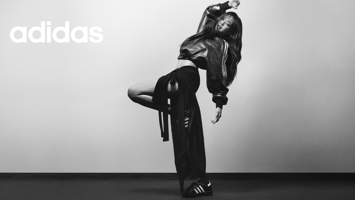 Global music icon JENNIE captured in a dynamic, artistic pose wearing a textured black adidas jacket, matching trousers, and the iconic Superstar sneakers.
