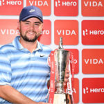 Alex Fitzpatrick smiling and holding the 2026 Hero Indian Open winner's trophy in front of a red Hero and Vida branding backdrop.