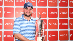Alex Fitzpatrick smiling and holding the 2026 Hero Indian Open winner's trophy in front of a red Hero and Vida branding ba...