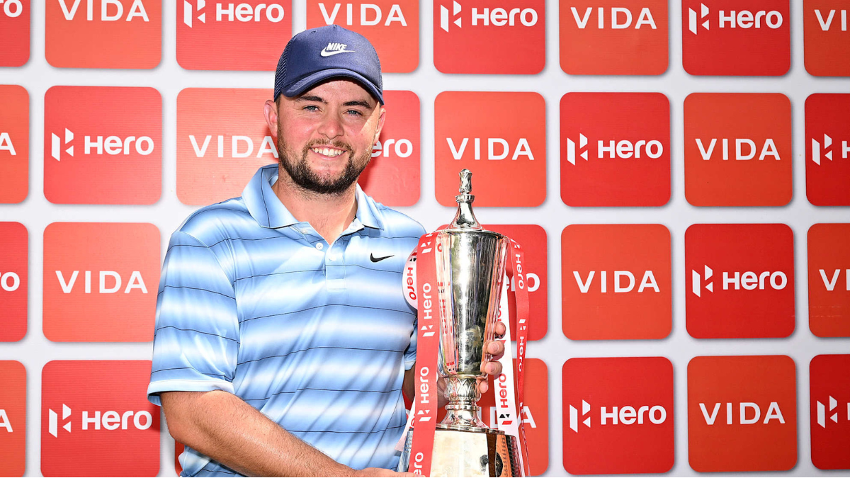 Alex Fitzpatrick smiling and holding the 2026 Hero Indian Open winner's trophy in front of a red Hero and Vida branding backdrop.