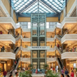 The sprawling multi-level atrium of Avenue Mall at Fortune City, featuring a central glass elevator, modern escalators, and premium storefronts under a large glass skylight.