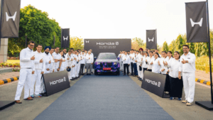 A large group of Honda Cars India employees giving thumbs-up gestures, standing on a red carpet in front of a blue-camoufl...