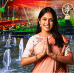 A welcoming woman in a peach saree performing a Namaste gesture in front of the illuminated Bharat Mandapam and Nataraja statue at night.