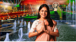 A welcoming woman in a peach saree performing a Namaste gesture in front of the illuminated Bharat Mandapam and Nataraja s...