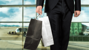 A traveller in a formal suit holding duty-free shopping bags at an airport terminal with a plane in the background, repres...
