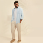 A mid-shot of Jasprit Bumrah smiling, featuring a light blue Uniqlo open linen shirt over a white inner tee against a neutral background.