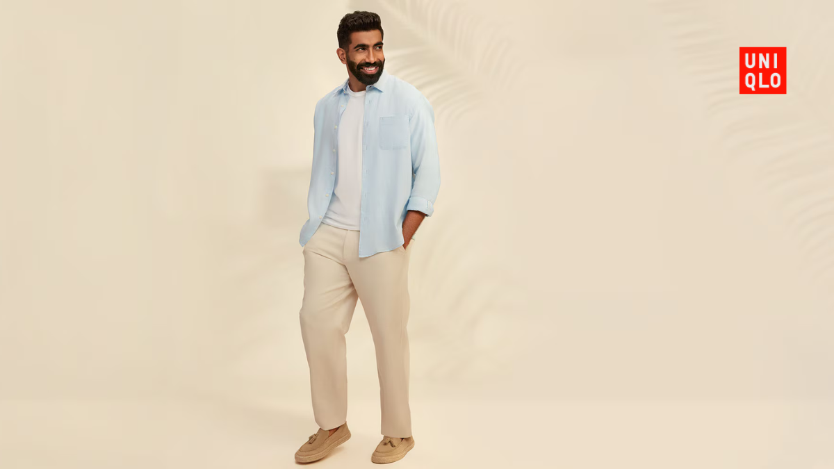 A mid-shot of Jasprit Bumrah smiling, featuring a light blue Uniqlo open linen shirt over a white inner tee against a neutral background.