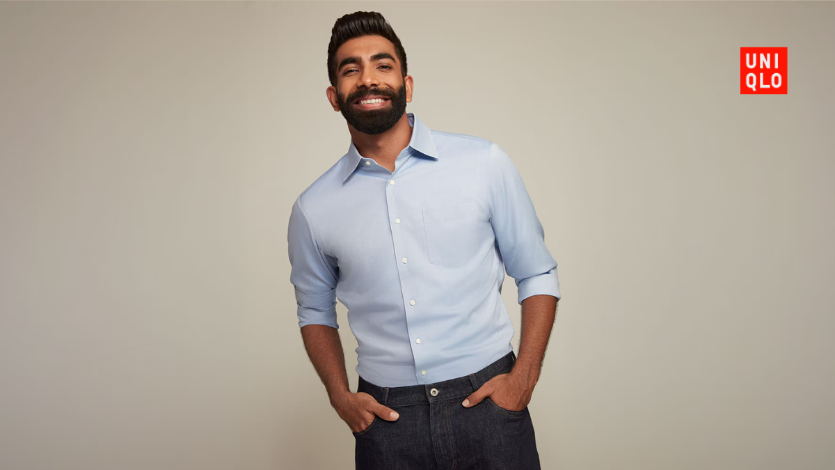A waist-up portrait of Jasprit Bumrah smiling in a tucked-in light blue Uniqlo button-down shirt paired with dark denim.