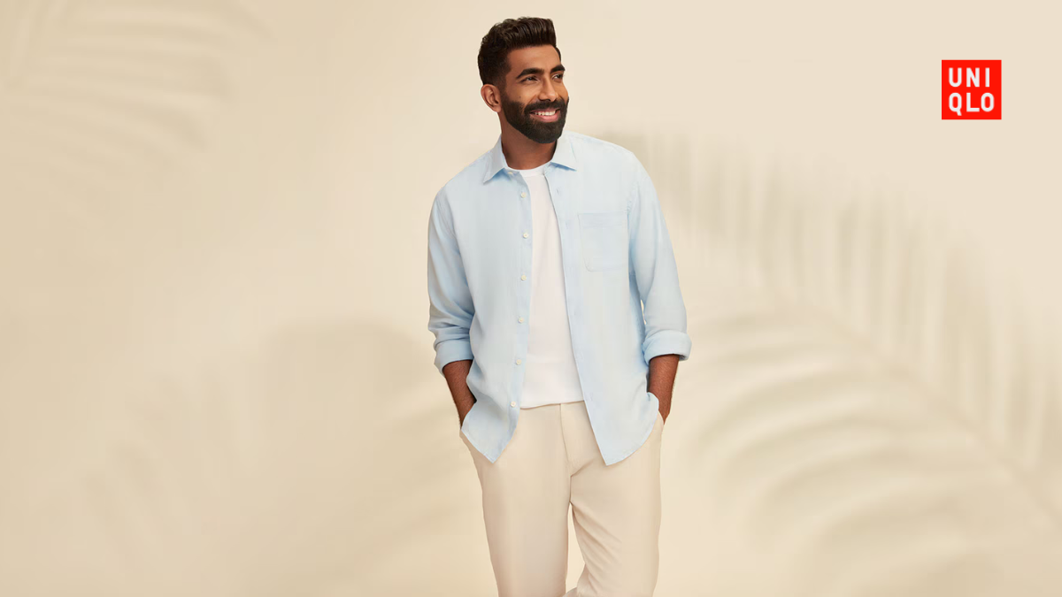 A mid-shot of Jasprit Bumrah smiling, featuring a light blue Uniqlo open linen shirt over a white inner tee against a neutral background.