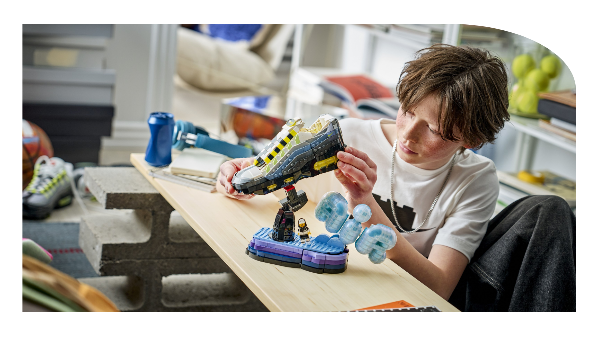 A wide top-down shot of a young boy in his personalized bedroom looking proudly at the completed LEGO Air Max 95 model on its display stand, surrounded by a skateboard, a basket of tennis balls, and another real Air Max sneaker.