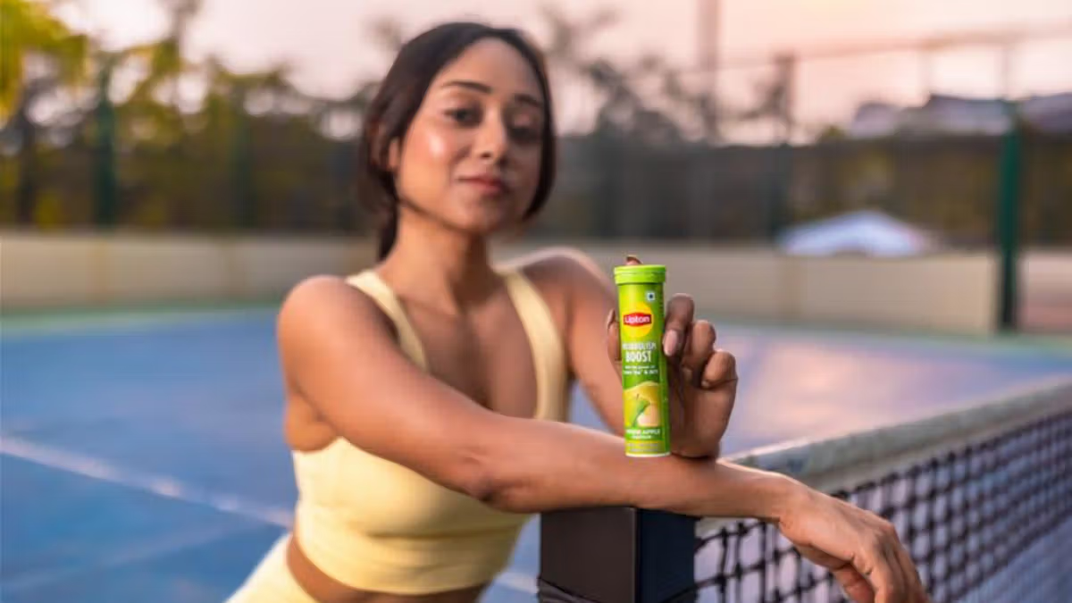 A woman in sports gear holding a Lipton Metabolism Boost Green Apple effervescent tube on a tennis court, representing a modern Indian wellness routine.