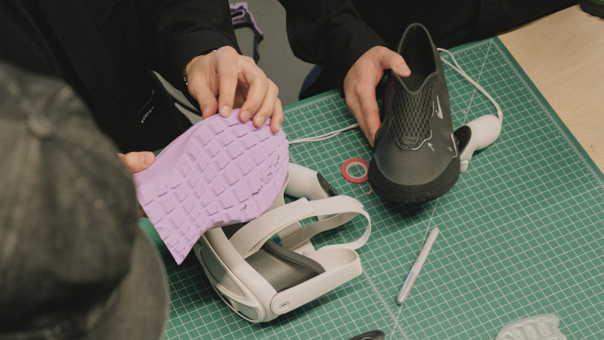 A designer holding a purple 3D-printed outsole next to a white VR headset and a black sneaker upper on a workbench.