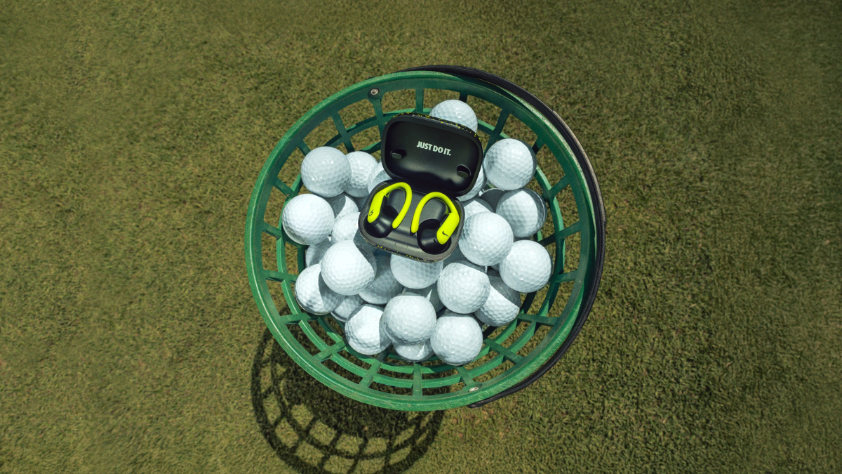Top-down view of the open Nike x Powerbeats Pro 2 charging case sitting inside a green basket of golf balls, revealing the 'Just Do It' interior branding.