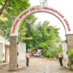 The main entrance arch of Nisargopchar Ashram in Uruli Kanchan, Pune, featuring signs in Marathi and English with security personnel at the gate.