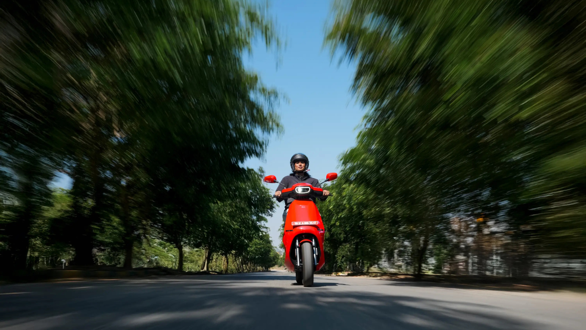 A rider on a red Ola S1 scooter moving forward on a tree-lined road with a dynamic motion blur effect.