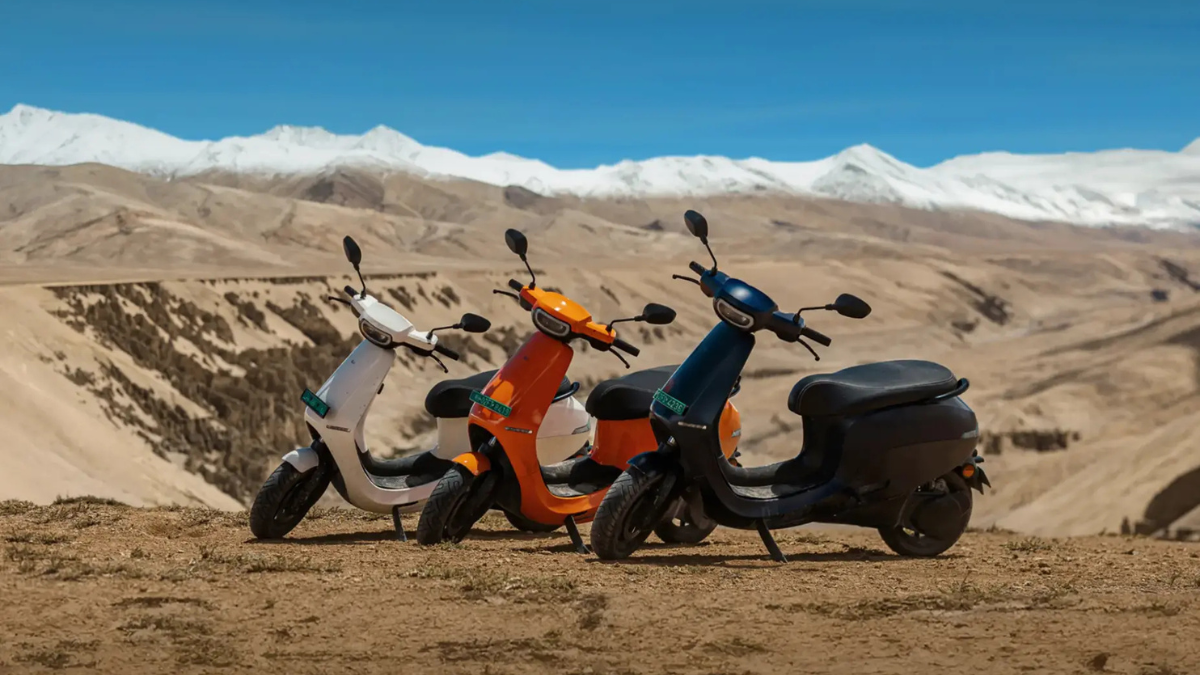 Three Ola S1 electric scooters in white, orange, and blue parked on a dirt trail with snow-capped mountains in the background.