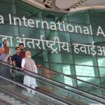 Prime Minister Narendra Modi and Uttar Pradesh officials on an escalator in front of the large "Noida International Airport" signage in English and Hindi.