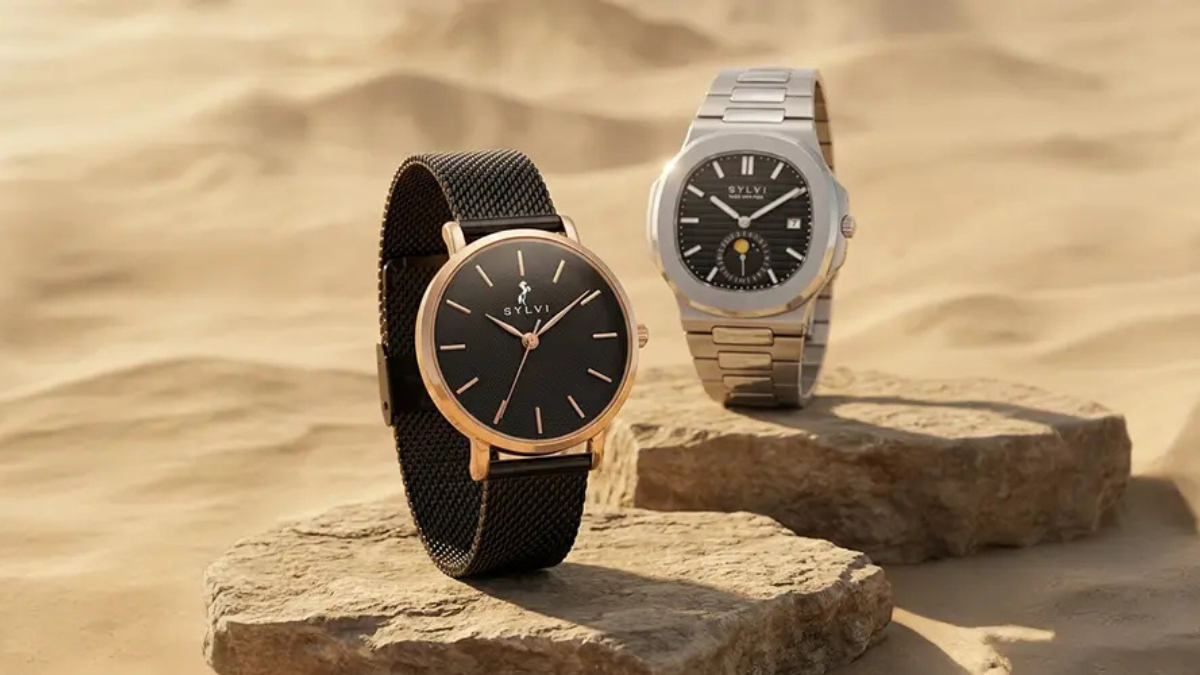 Sylvi Summer Sale 2026: Upgrade Your Wrist Game with Trending Watches for Men and Women (Up to 18% Off) 2 Product photography of two Sylvi watches. One watch on the left is a round rose gold case with a black dial, gold indices and hands, and a black metal mesh strap. It is on a small rough rock. The other watch on the right is a silver-toned metal case with a distinct octagonal bezel, an integrated silver bracelet, a black waffle-patterned dial, and a moon phase complication at 6 o'clock. It is on a separate small rough rock. Both watches are in the center, resting on the rock, with warm sunlight illuminating them and casting shadows over the desert sand dunes and rough texture below and behind them.