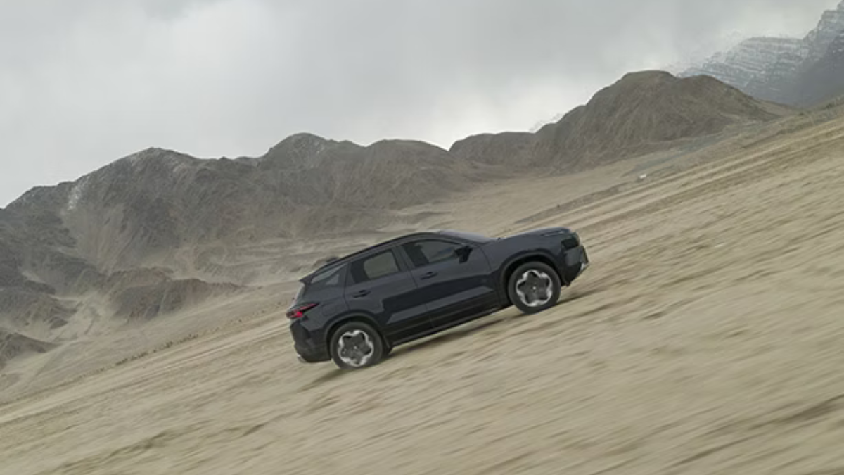 Side view of a dark Tata Harrier.ev driving across a sandy desert plain with mountains in the background.