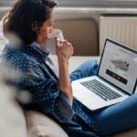 A freelancer drinking coffee while viewing a "Your Cart" page on a laptop, showing a global sneaker purchase in USD with a Mastercard checkout option.