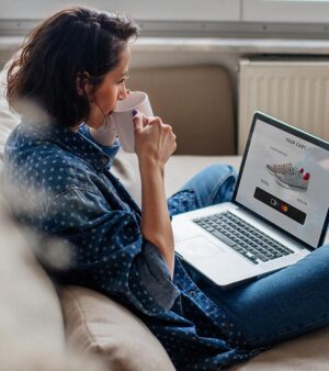 A freelancer drinking coffee while viewing a "Your Cart" page on a laptop, showing a global sneaker purchase in USD with a...