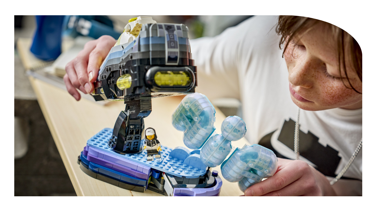A young boy with freckles and a necklace actively assembling or adjusting the completed 1,213-piece LEGO Air Max 95 model on a detailed purple and blue display base, featuring the translucent 'P' logo structure and customizable minifigure in a white cap.