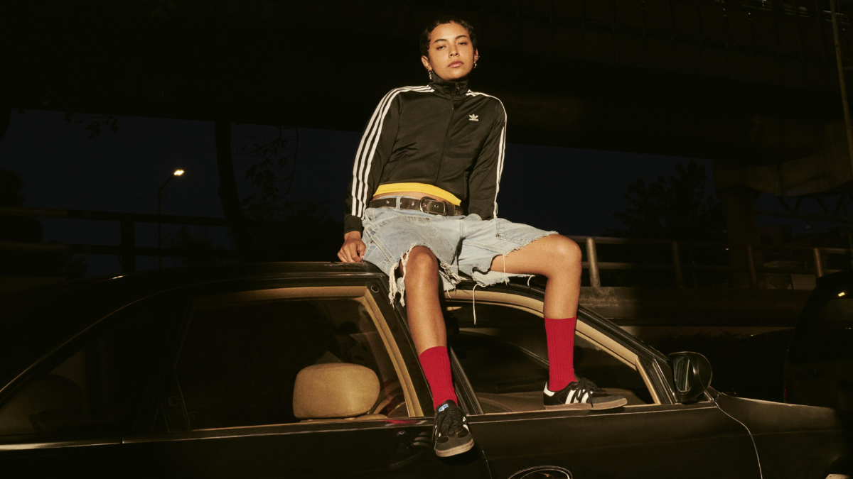 A person sitting on top of a black car at night wearing an adidas Firebird track jacket, denim shorts, and black Sambas.