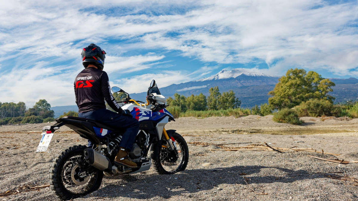 Rear-quarter view of a rider sitting on a BMW F 450 GS motorcycle on a sandy beach, looking towards a distant snow-capped mountain