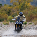 Head-on view of a rider in full gear on a BMW F 450 GS motorcycle crossing a shallow river, creating water splashes