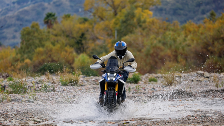 Head-on view of a rider in full gear on a BMW F 450 GS motorcycle crossing a shallow river, creating water splashes