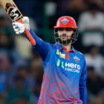 A Delhi Capitals cricket player celebrating a milestone on the field, raising an SS cricket bat to acknowledge the crowd. He wears a red helmet and blue/orange team jersey with 'Hero FINCORP' branding during a TATA IPL match.