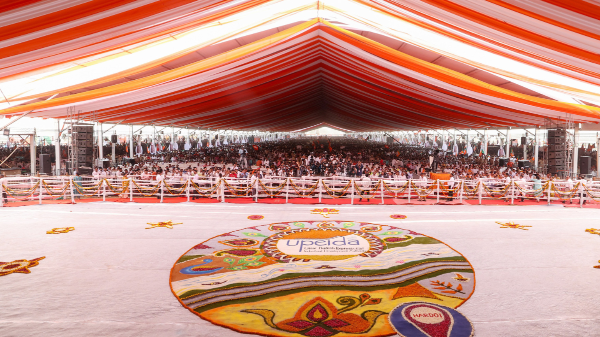 Massive crowd seated under an orange and white tent behind a large UPEIDA floor rangoli at the Ganga Expressway launch.