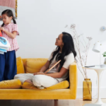 A mother and child on a yellow sofa, interacting near a Google Nest smart display and speaker.