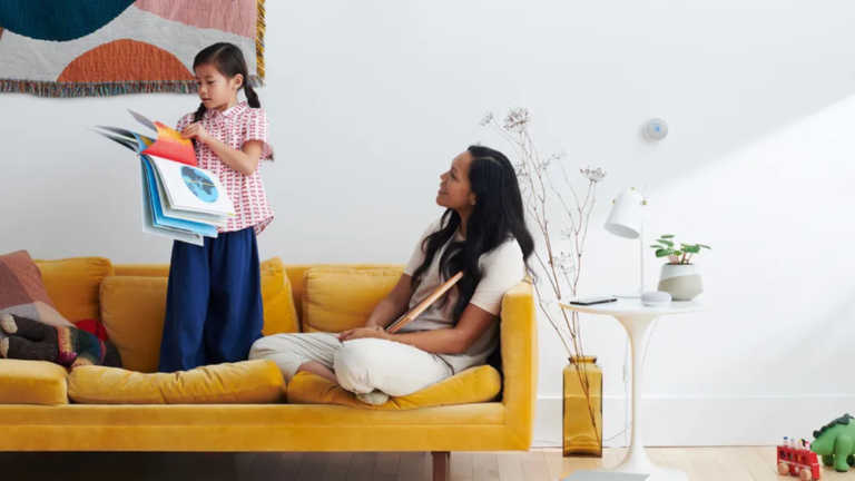 A mother and child on a yellow sofa, interacting near a Google Nest smart display and speaker.