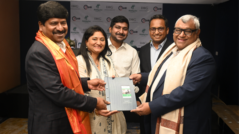 Five individuals exchanging a blue IHG folder in front of an IHG and TGH&R banner during a signing in India.