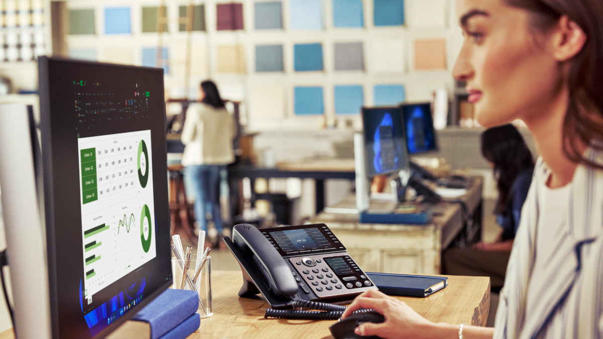 A professional woman working at a desk in a creative office environment with an HP Poly Edge V Series desk phone and data ...