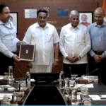 Eight Indian officials in a boardroom exchanging signed government agreements in brown folders for Jal Jeevan Mission.