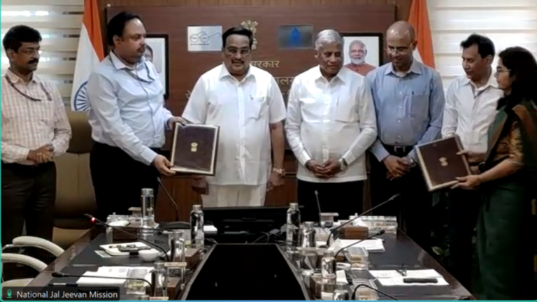 Eight Indian officials in a boardroom exchanging signed government agreements in brown folders for Jal Jeevan Mission.