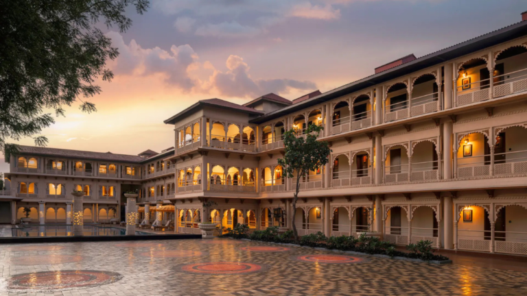 A wide-angle, detailed dusk photograph of a multi-story traditional Indian palace hotel facade surrounding an enclosed cou...