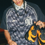 A man with braided hair wearing a patterned Nike Air Max 90 button-down shirt holding a pair of vibrant orange and black H...